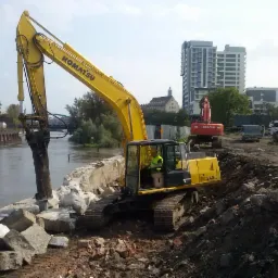 Żółta koparka Komatsu z hydraulicznym młotem kruszy betonowe nabrzeże rzeki, w tle budynki miejskie i druga koparka.