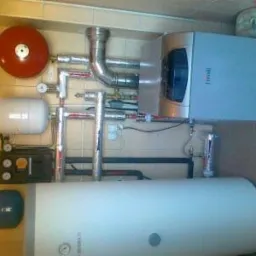 Nowoczesny piec gazowy marki Ferroli z podłączonym zbiornikiem ciepłej wody użytkowej, widoczne rury instalacyjne oraz elementy sterujące, zamontowany w pomieszczeniu z wykończeniem w płytkach.