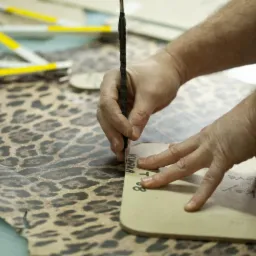 Ręczne wycinanie szablonu z materiału o wzorze panterki przy pomocy ołówka i szablonu z napisem 'KLAPKA T. 8'.