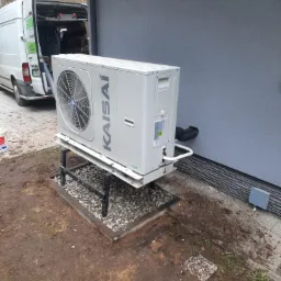 Biała pompa ciepła marki Kaisai zamontowana na metalowej podstawie przy szarej ścianie budynku, z widocznym samochodem dostawczym firmy instalacyjnej w tle.