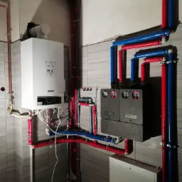 Nowoczesny, wiszący kocioł gazowy z podłączonymi rurami grzewczymi w kolorach czerwonym i niebieskim, zainstalowany w pomieszczeniu z szarą, kafelkową ścianą.