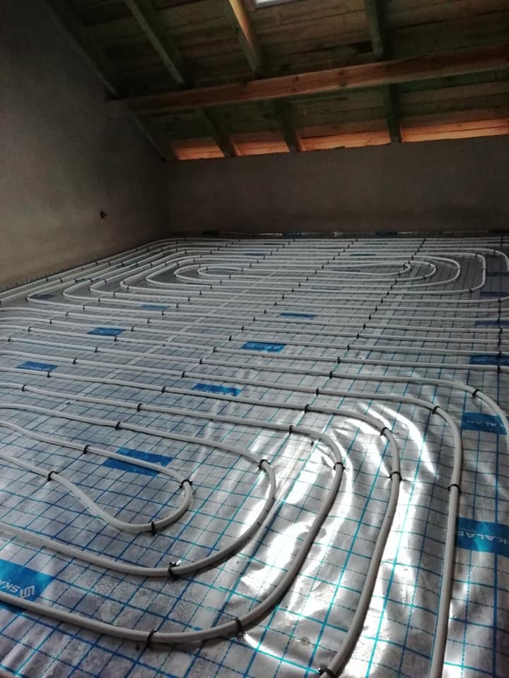 Białe rurki ogrzewania podłogowego ułożone spiralnie na izolacji z niebieską siatką, widok z góry, poddasze z drewnianą konstrukcją dachu w tle.