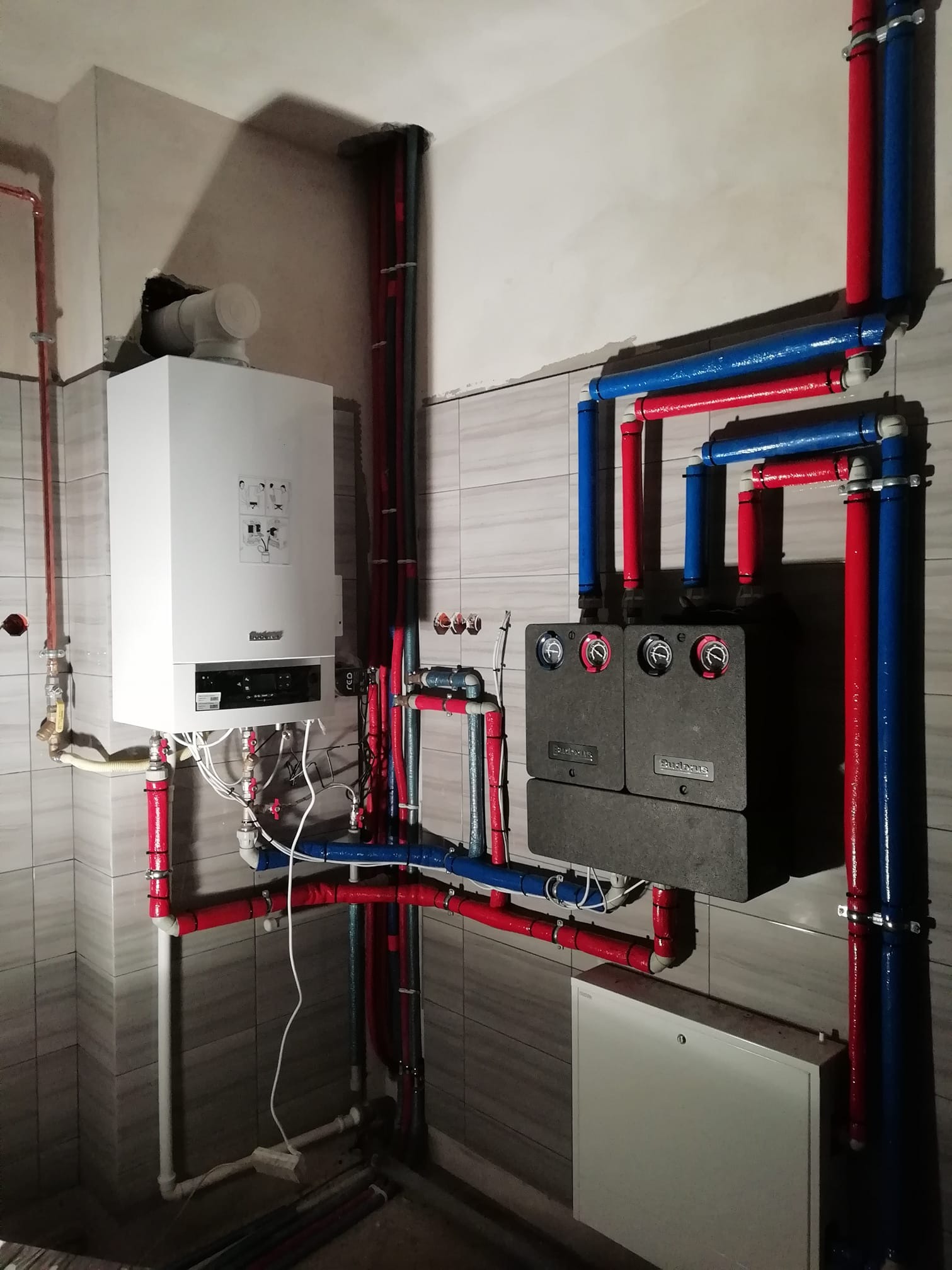 Nowoczesny, wiszący kocioł gazowy z podłączonymi rurami grzewczymi w kolorach czerwonym i niebieskim, zainstalowany w pomieszczeniu z szarą, kafelkową ścianą.