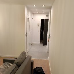 Baranski - Jasne wnętrze mieszkania po remoncie: widok z salonu na korytarz z białymi ścianami, szarą podłogą i wbudowanymi punktowymi lampami sufitowymi, częściowo widoczna szara sofa z folią ochronną.