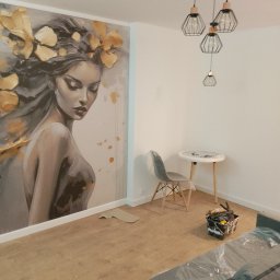 Baranski - Pokój w trakcie prac, ściana z dużym malowidłem kobiety z kwiatami, zabezpieczona folią sofa, wiadro z narzędziami malarskimi, mały stolik i designerska lampa wisząca.