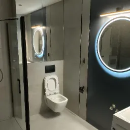 Nowoczesna łazienka z podwieszaną miską WC, lustrem z podświetleniem LED i kabiną prysznicową z czarnymi profilami, wykończona w szarościach z akcentem ciemnego granatu.