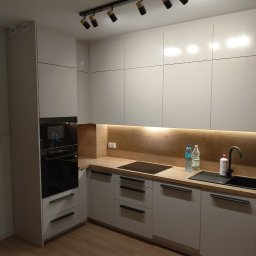 Baranski - Nowoczesna kuchnia z białymi, matowymi szafkami, drewnianym blatem i czarnymi uchwytami. Nad blatem zamontowane oświetlenie LED. Na blacie widoczne butelki z wodą i płyn do naczyń. Dodatkowe...