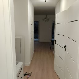 Baranski - Korytarz po remoncie z białymi drzwiami z poziomymi frezami, jasną podłogą i widocznymi narzędziami do montażu listew przy podłodze.