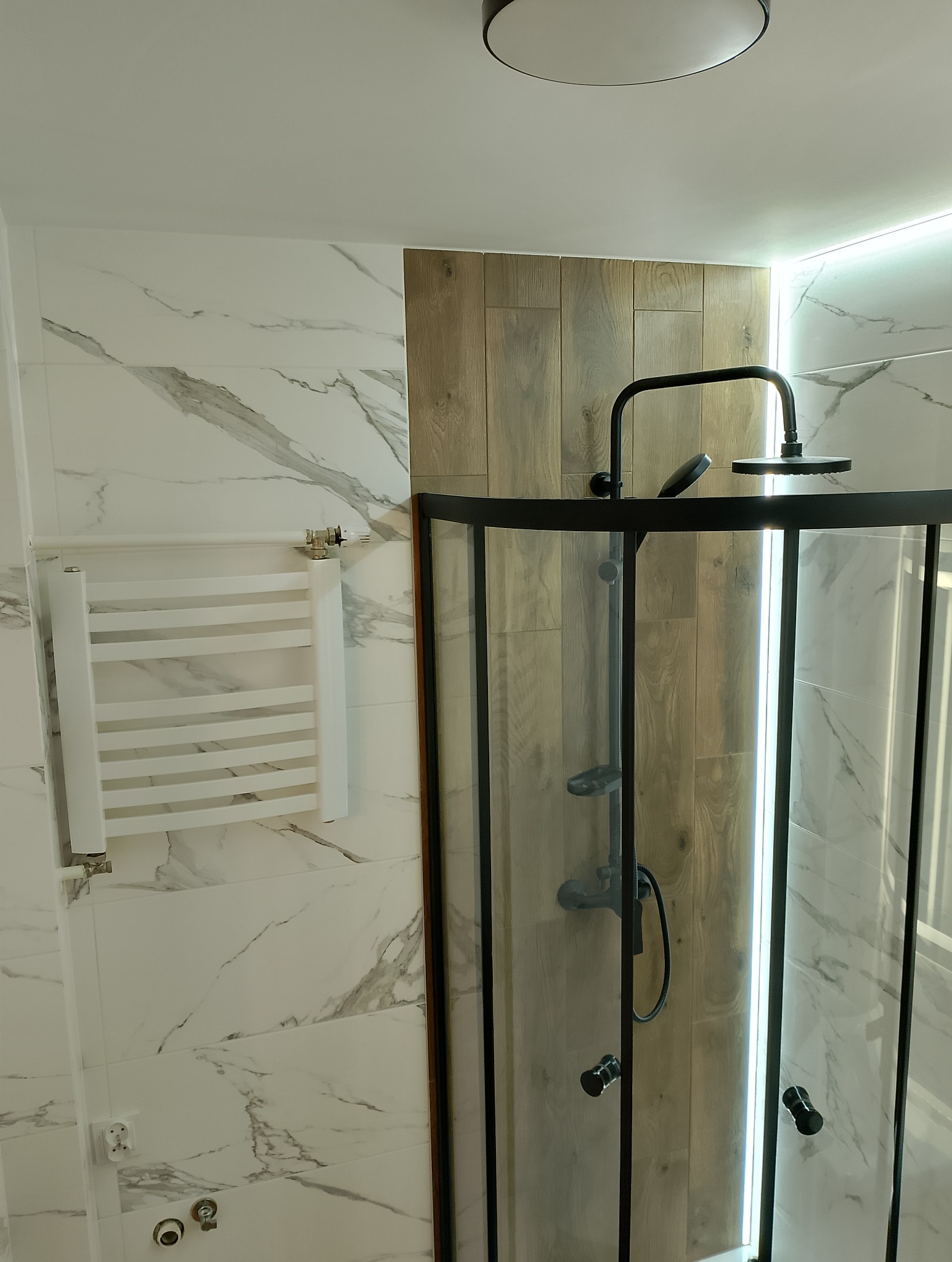 Nowoczesna kabina prysznicowa z czarną armaturą, drewnopodobne panele na ścianie, białe marmurowe płytki i grzejnik drabinkowy w łazience z oświetleniem LED.