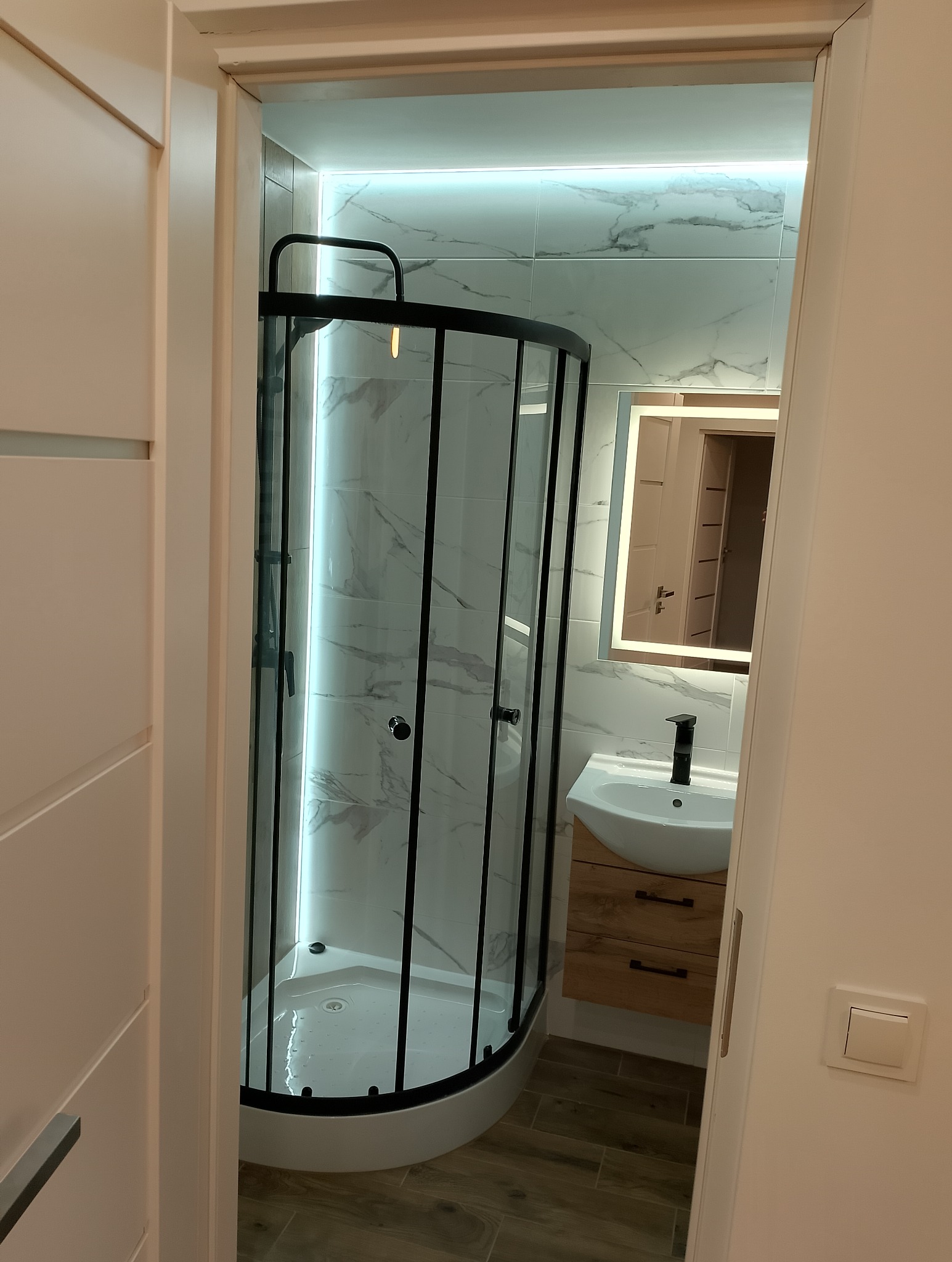 Nowoczesna łazienka z kabiną prysznicową narożną z czarnymi ramami, umywalką nablatową na drewnianej szafce i podświetlanym lustrem, wykończona białymi płytkami imitującymi marmur.