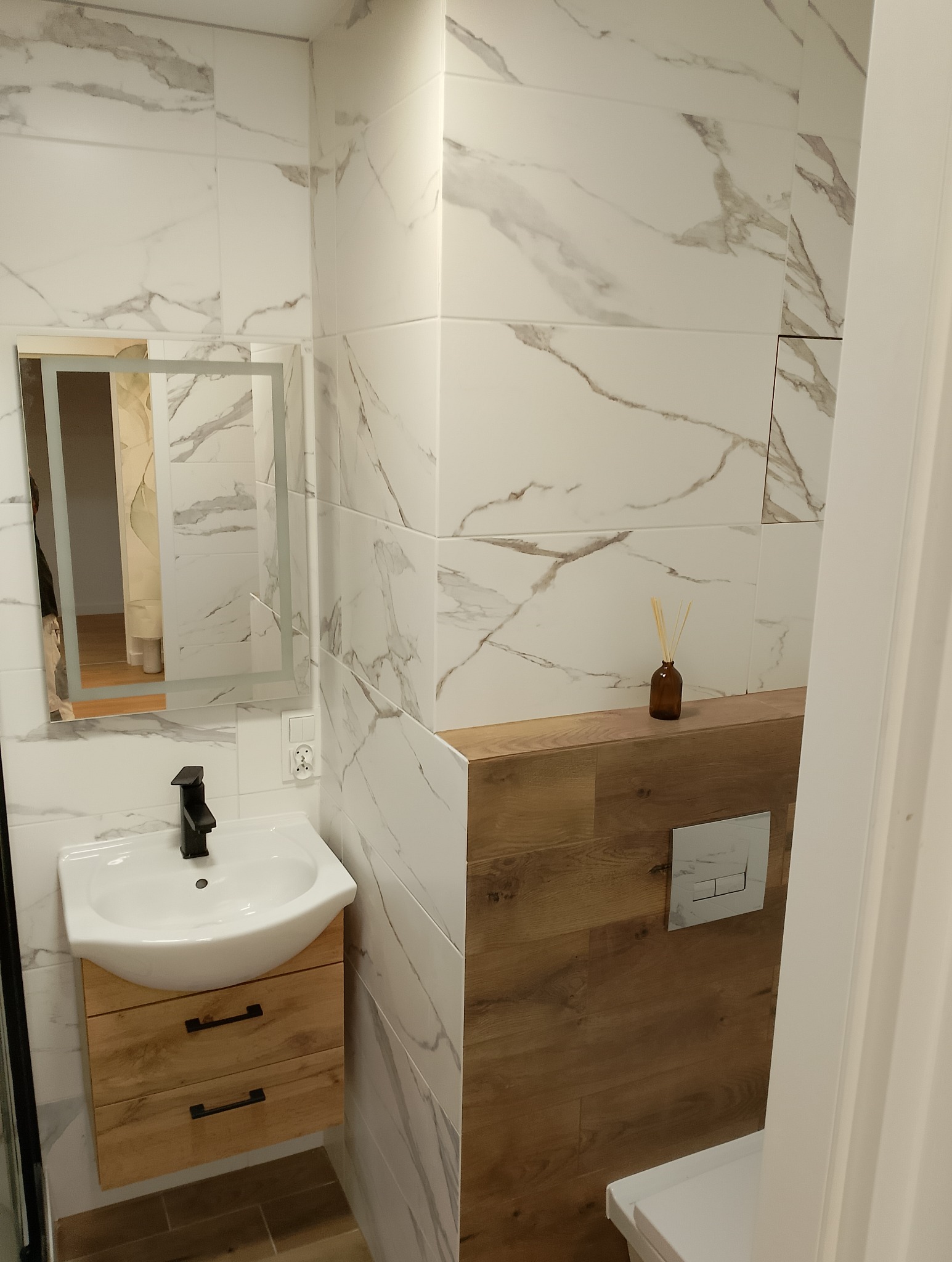 Nowoczesna łazienka z białymi płytkami imitującymi marmur, drewnianą szafką pod umywalką z czarną armaturą i podświetlanym lustrem. Uwagę zwraca drewniana obudowa stelaża WC z dyskretnym...