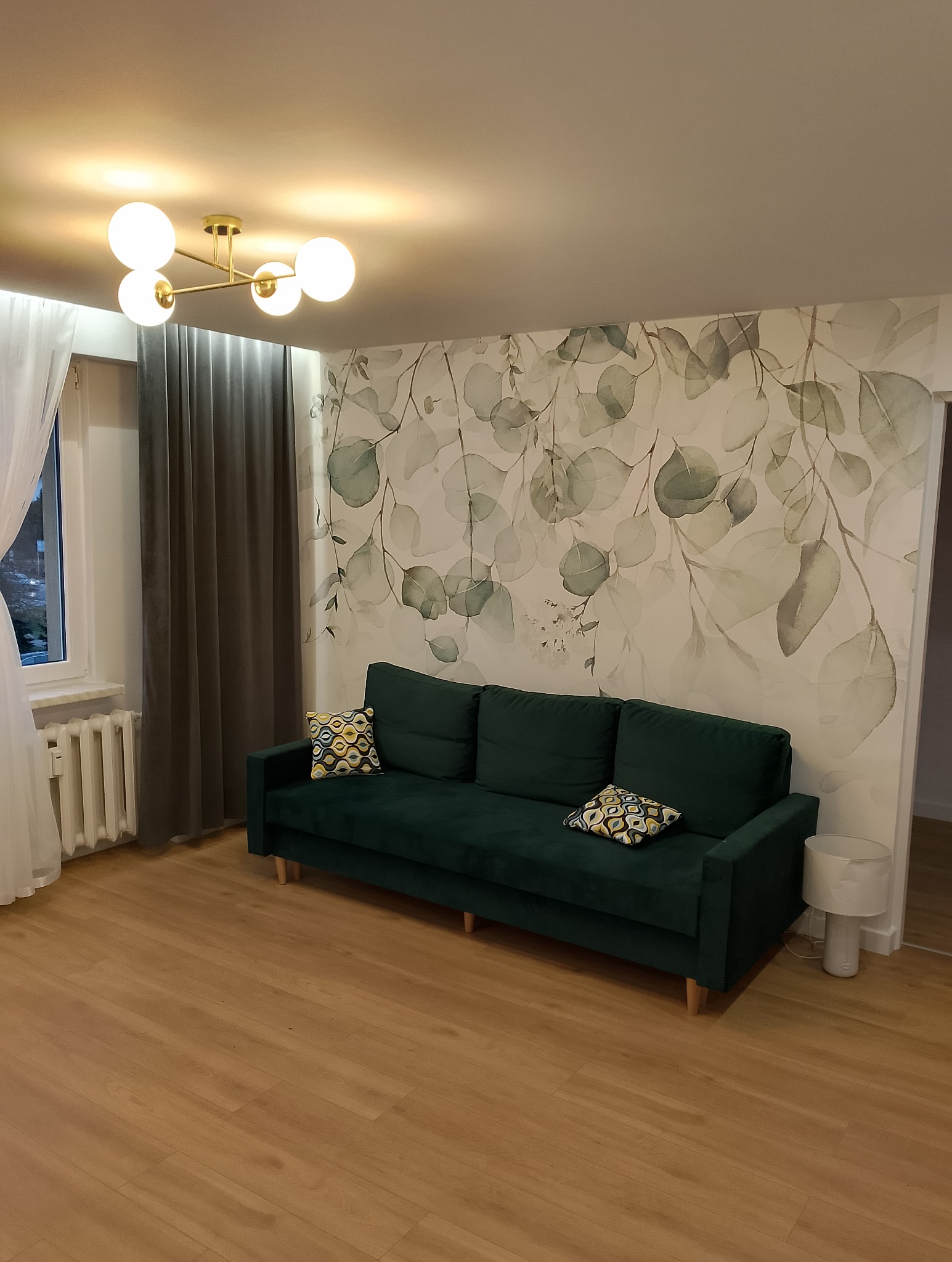 Pokój z zieloną sofą na tle ściany z motywem liści, drewniana podłoga, okno z zasłonami i nowoczesna lampa sufitowa.