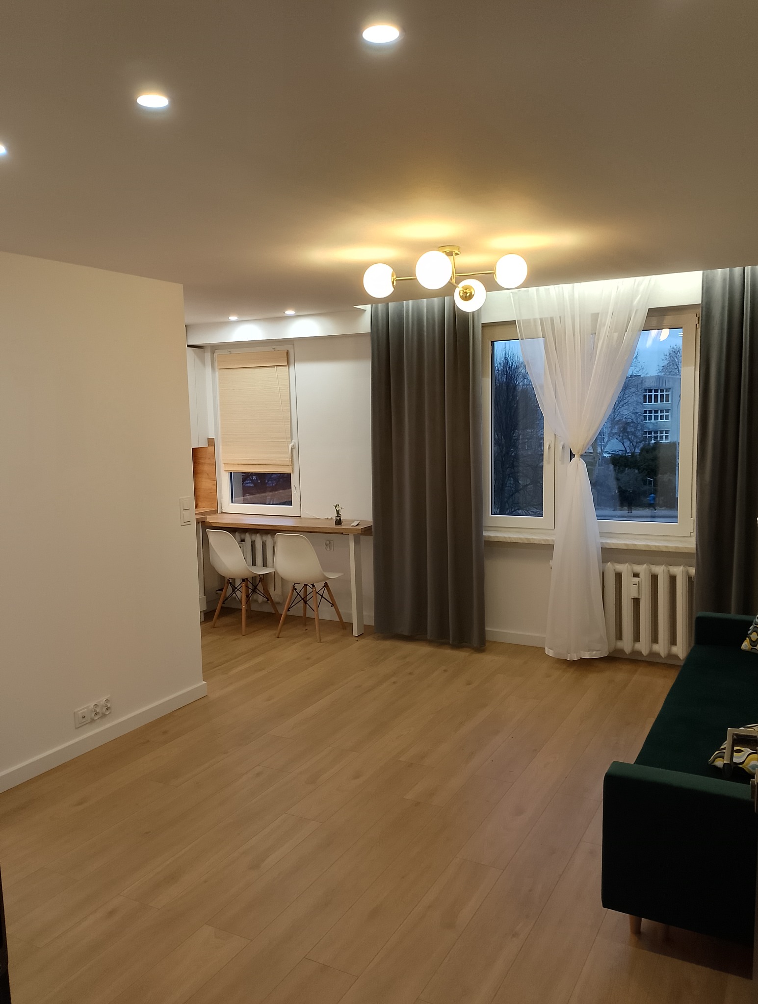 Jasne, przestronne wnętrze pokoju po remoncie: drewniana podłoga, białe ściany, okno z szarymi i białymi zasłonami, designerska lampa sufitowa, biurko przy oknie z drewnianym blatem i białymi...