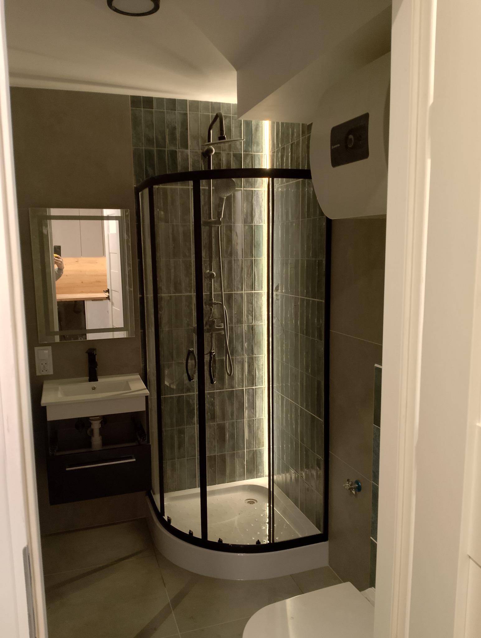 Nowoczesna kabina prysznicowa z czarnymi profilami i zielonymi płytkami w wyremontowanej łazience, widok zza framugi drzwi.