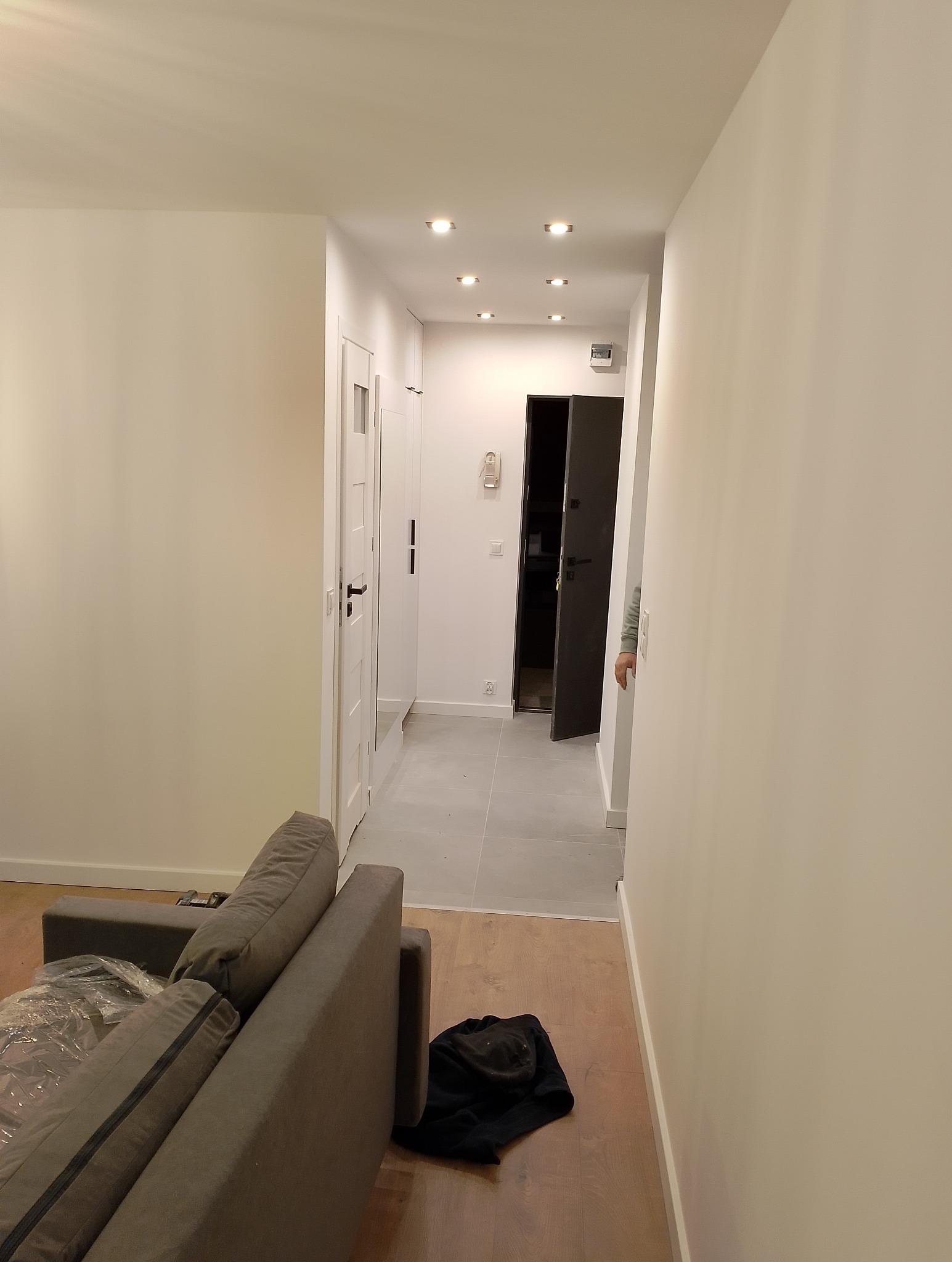 Jasne wnętrze mieszkania po remoncie: widok z salonu na korytarz z białymi ścianami, szarą podłogą i wbudowanymi punktowymi lampami sufitowymi, częściowo widoczna szara sofa z folią ochronną.