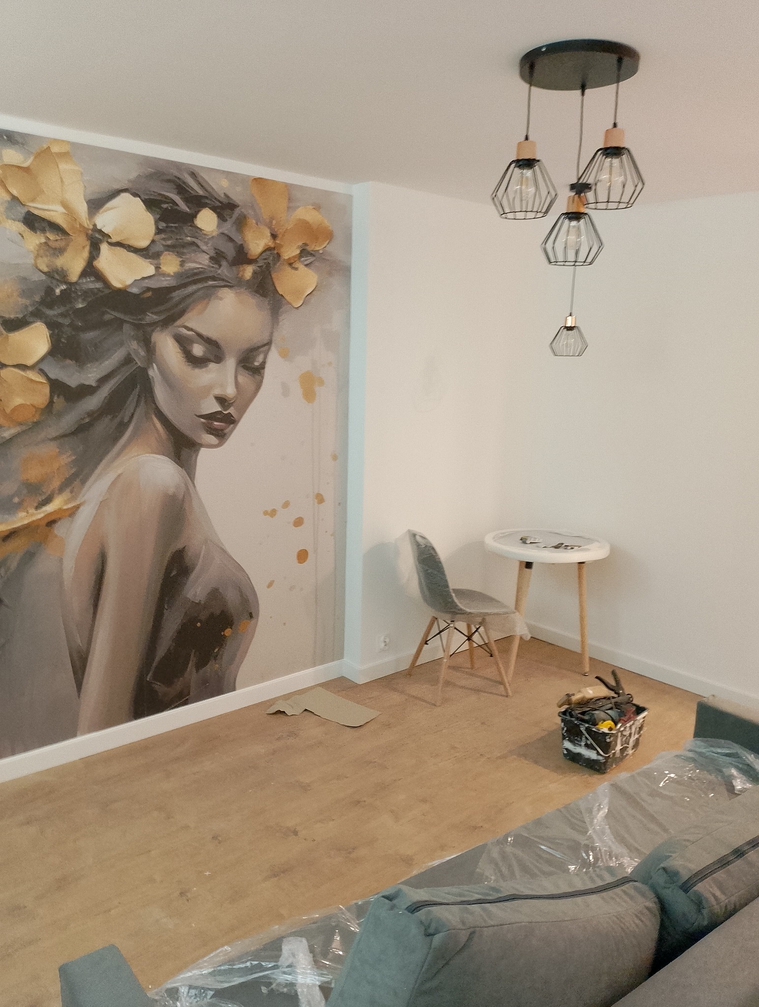 Pokój w trakcie prac, ściana z dużym malowidłem kobiety z kwiatami, zabezpieczona folią sofa, wiadro z narzędziami malarskimi, mały stolik i designerska lampa wisząca.