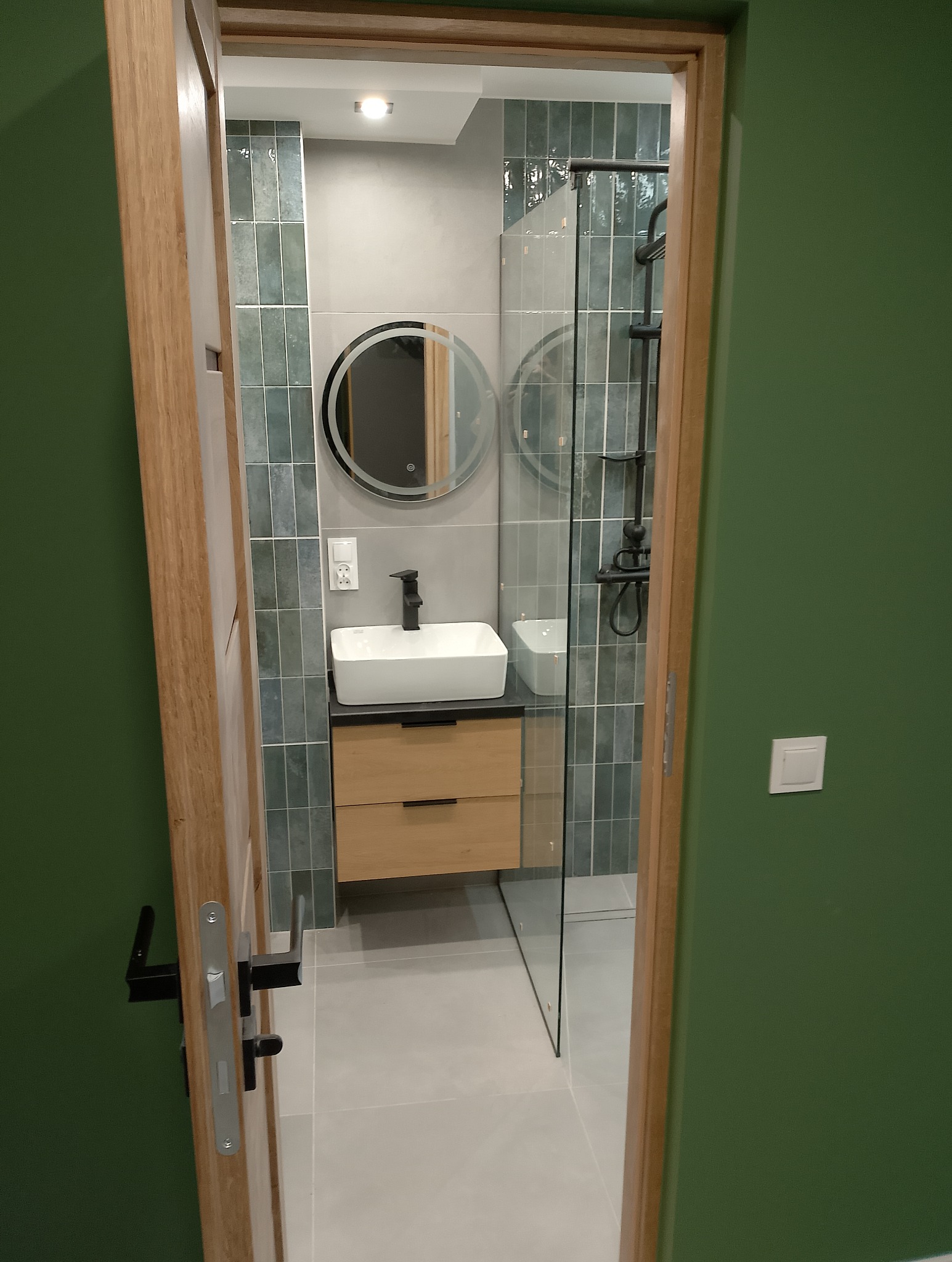 Widok na wykończoną łazienkę z kabiną prysznicową typu walk-in, umywalką nablatową na szafce z drewnianymi frontami, okrągłym lustrem z podświetleniem LED i płytkami w odcieniach szarości i zieleni...