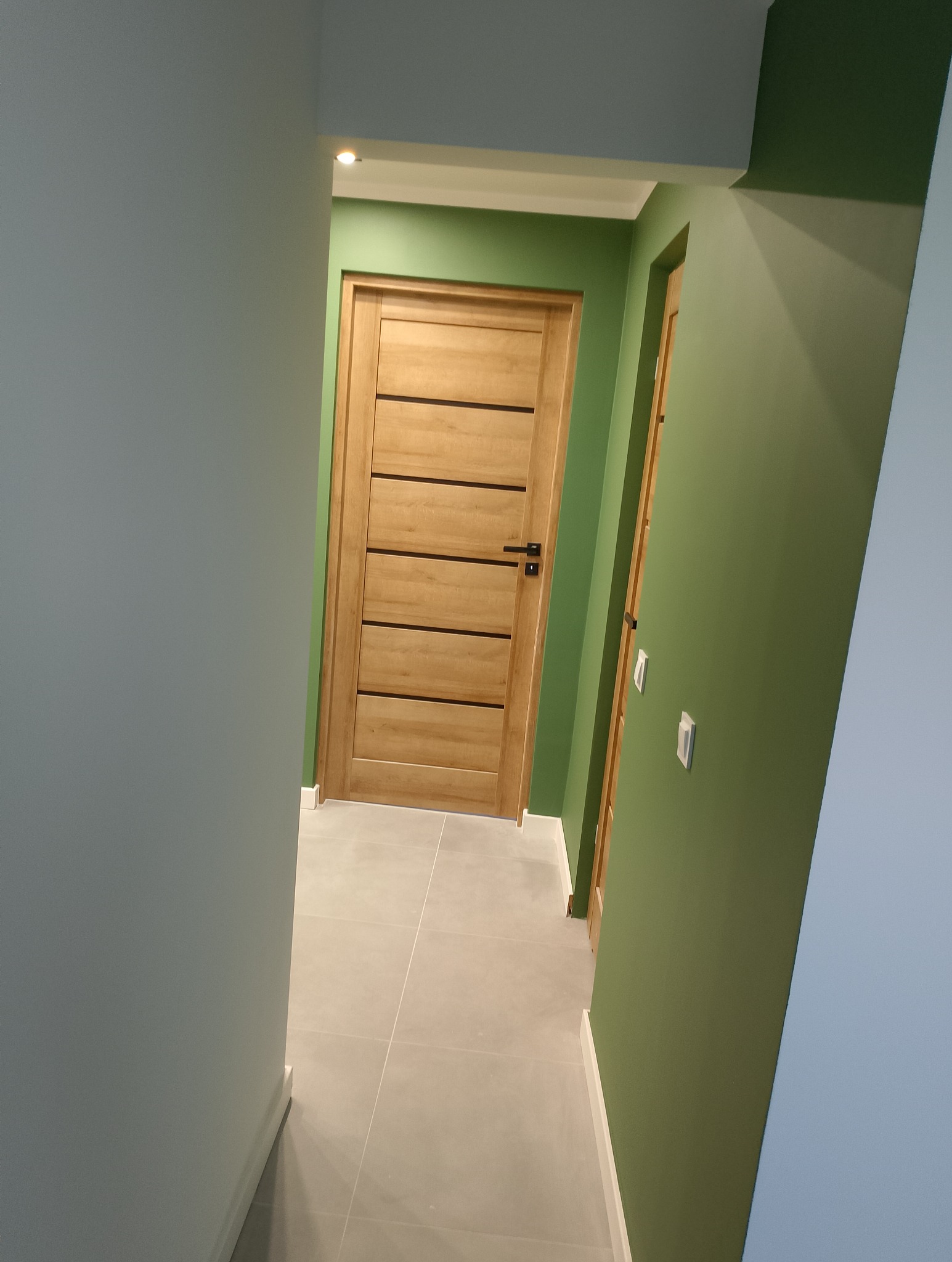 Korytarz z drewnianymi drzwiami z poziomymi wstawkami, pomalowany na zielono, z szarą podłogą z płytek i białymi listwami przypodłogowymi.
