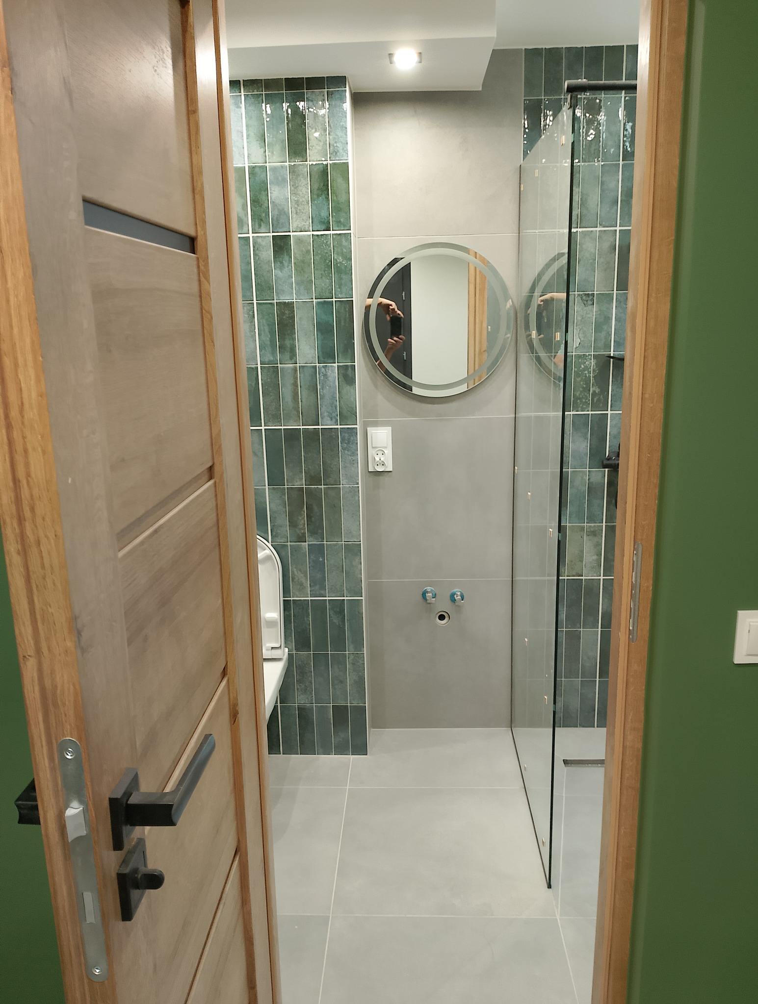 Nowoczesna łazienka z kabiną prysznicową typu walk-in, wykończona zieloną, pionową glazurą, widoczna przez drewniane drzwi z czarną klamką.
