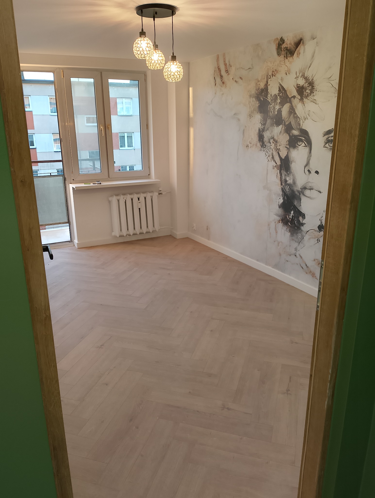 Wykończony pokój z parkietem ułożonym w jodełkę, dekoracyjną tapetą z motywem twarzy, trzema szklanymi lampami wiszącymi i białym grzejnikiem pod oknem.