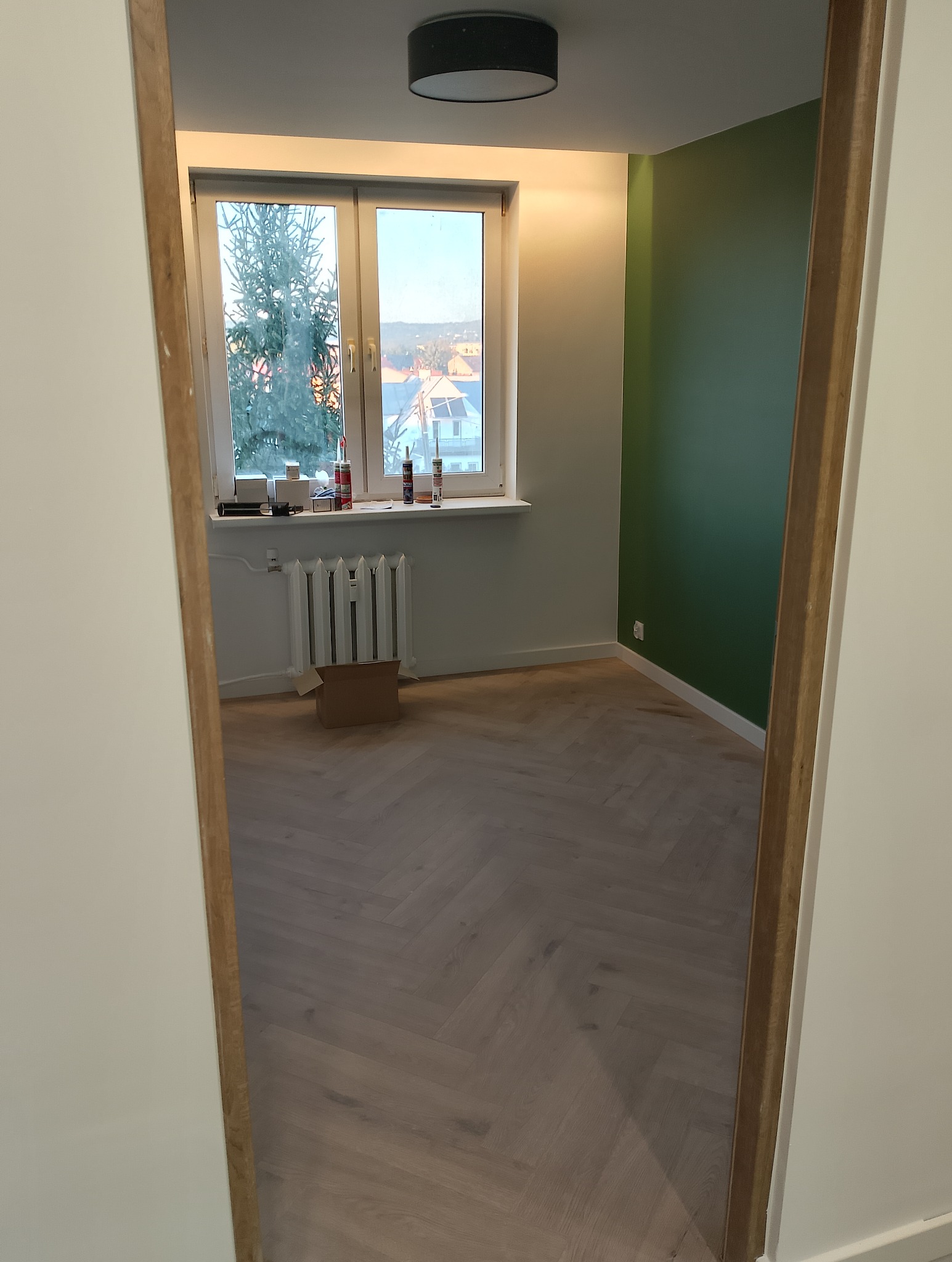 Pokój po remoncie z nową podłogą ułożoną w jodełkę, świeżo pomalowaną zieloną ścianą i grzejnikiem pod oknem z narzędziami na parapecie.