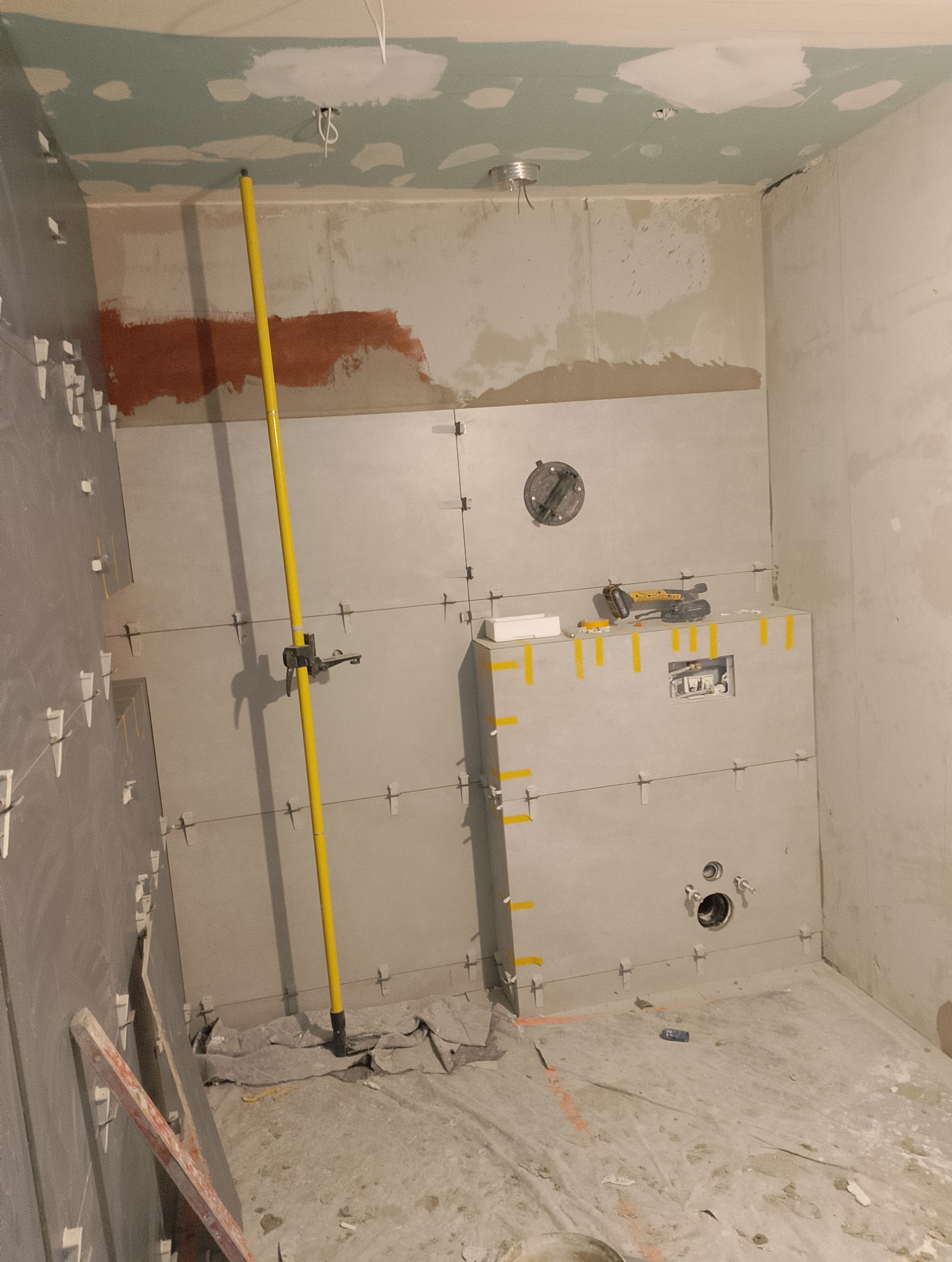 Łazienka w trakcie montażu płytek: szare płytki ułożone do połowy wysokości ścian, żółty pionownik, narzędzia glazurnicze na półce, widoczne instalacje wodne i elektryczne, surowy sufit...
