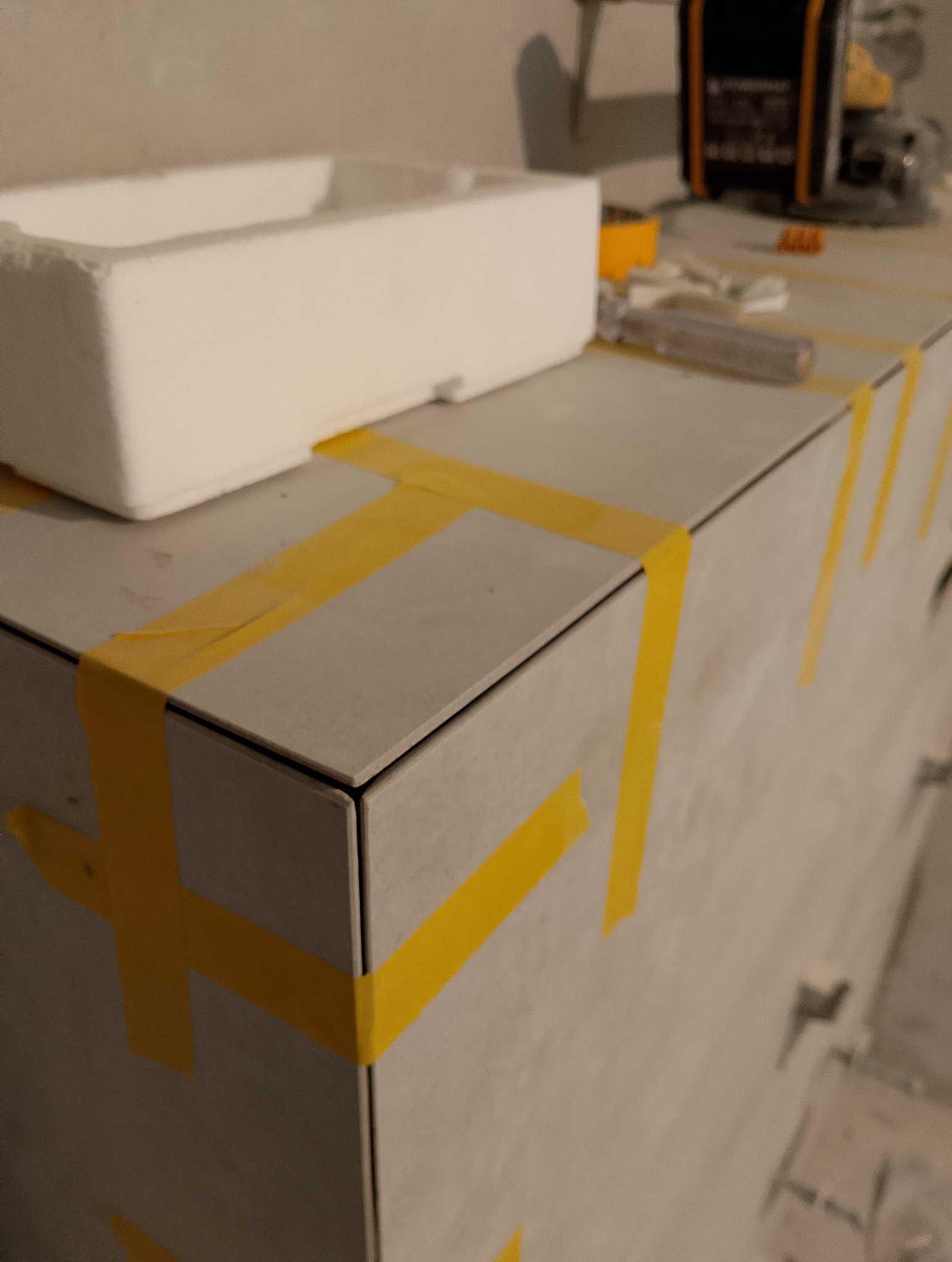 Szare płytki przyklejone do zabudowy, zabezpieczone żółtą taśmą malarską, na górze biały styropianowy pojemnik i narzędzia w tle.