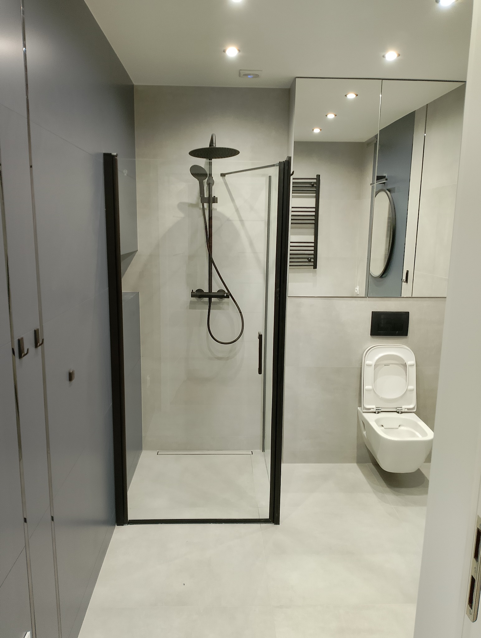 Nowoczesna łazienka z kabiną prysznicową typu walk-in z czarnymi ramami, wiszącą toaletą i szafkami w odcieniach szarości, oświetlona punktowymi lampami sufitowymi.