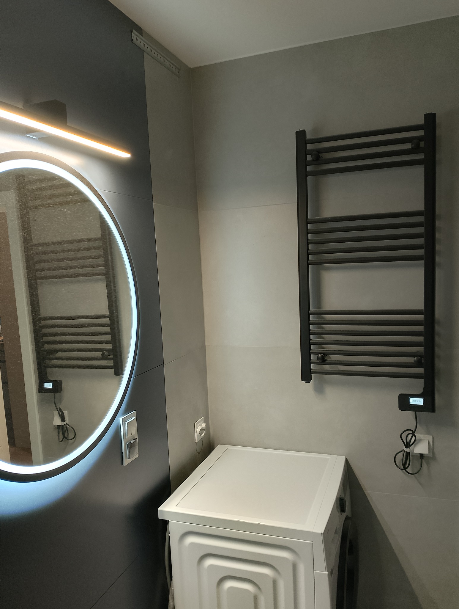 Nowoczesna łazienka w szarościach z okrągłym lustrem z podświetleniem LED, grzejnikiem drabinkowym w kolorze antracytowym i białą pralką.