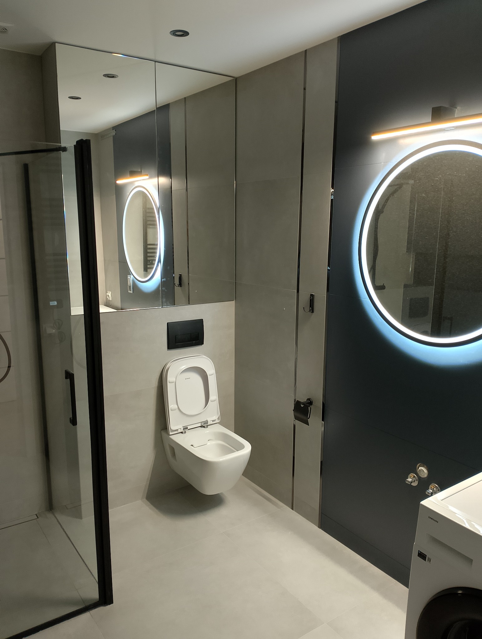 Nowoczesna łazienka z podwieszaną miską WC, lustrem z podświetleniem LED i kabiną prysznicową z czarnymi profilami, wykończona w szarościach z akcentem ciemnego granatu.