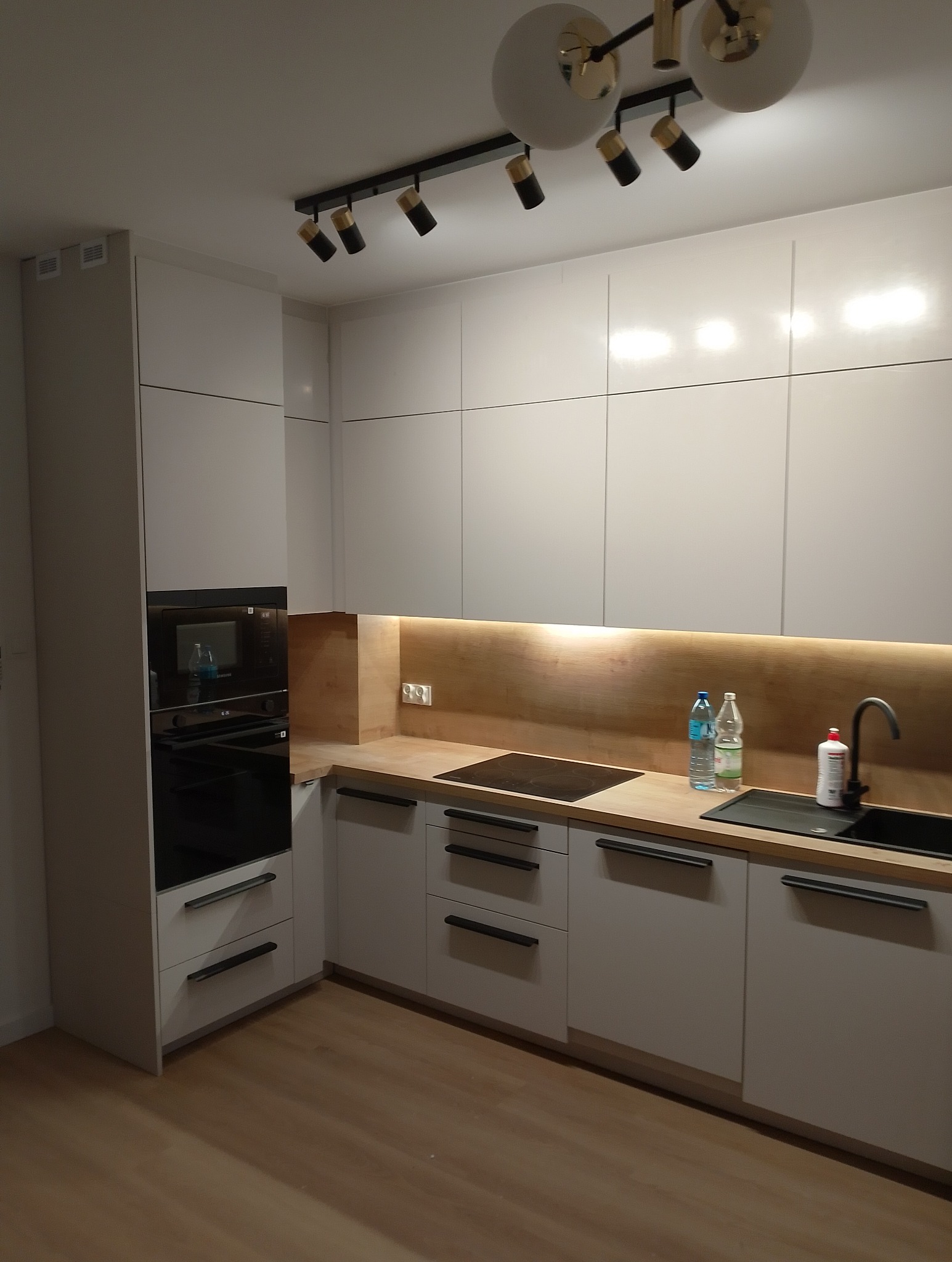 Nowoczesna kuchnia z białymi, matowymi szafkami, drewnianym blatem i czarnymi uchwytami. Nad blatem zamontowane oświetlenie LED. Na blacie widoczne butelki z wodą i płyn do naczyń. Dodatkowe...