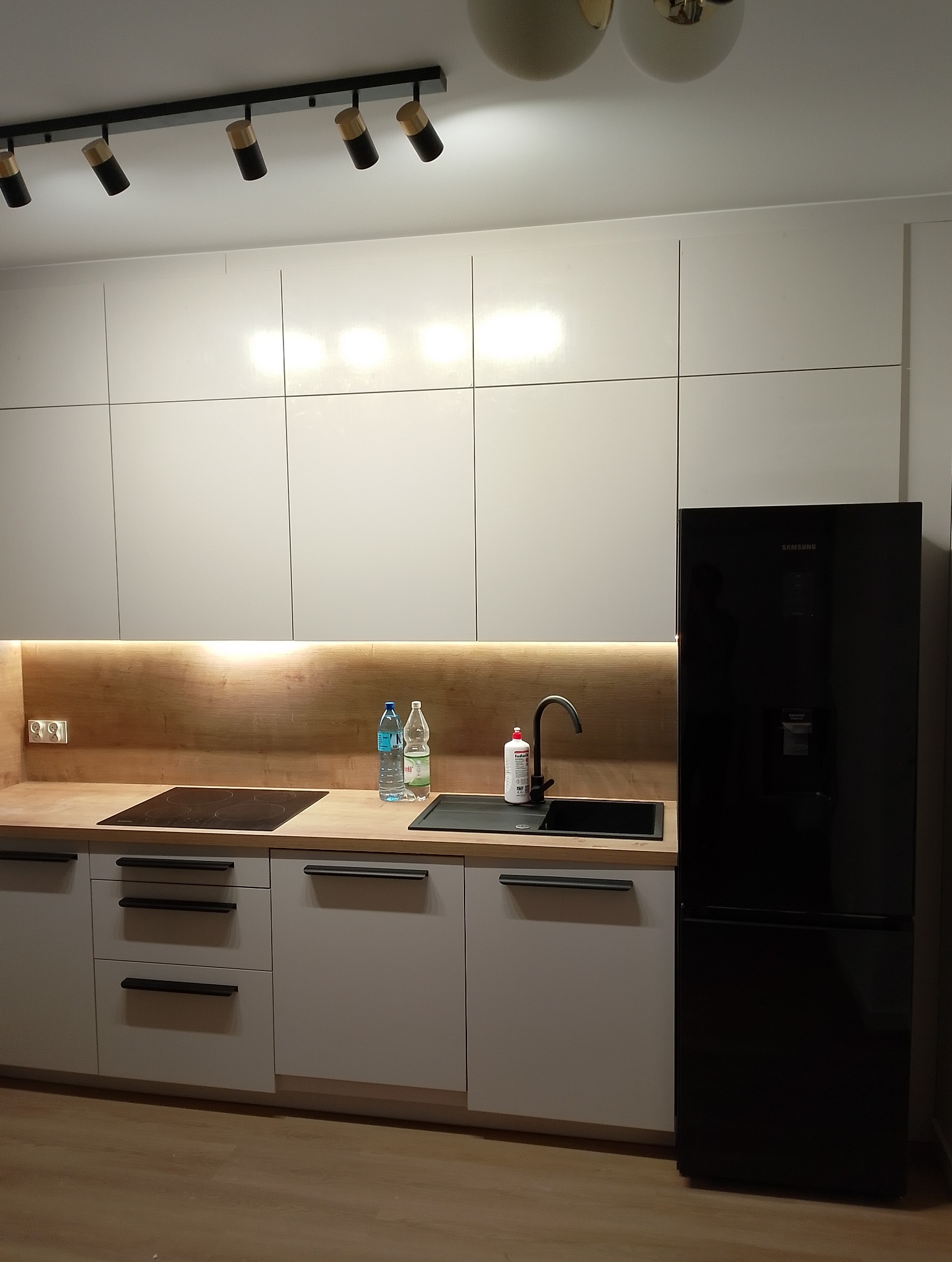 Nowoczesna kuchnia z białymi szafkami, drewnianym blatem, czarną armaturą i lodówką Samsung, oświetlona listwą LED pod szafkami wiszącymi i reflektorami na suficie.