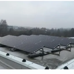 Rzędy paneli fotowoltaicznych zamontowanych na płaskim dachu budynku w pochmurny dzień, widoczne elementy mocujące i krajobraz w tle.