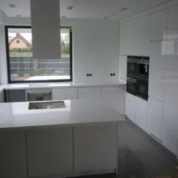 Nowoczesna, biała kuchnia z wyspą i dużym oknem, jeszcze w trakcie montażu, widoczne zabezpieczenia na podłodze.