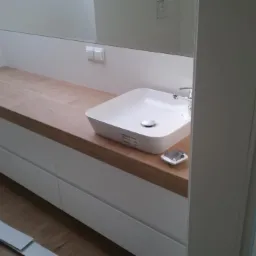 Nowoczesna łazienka: biała umywalka nablatowa na drewnianym blacie z białymi szufladami, minimalistyczny design, widoczne fragmenty listew przypodłogowych na podłodze.