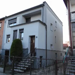 Odnowiona elewacja dwukondygnacyjnego domu z numerem 14, szara kolorystyka, nowoczesny design, widok z poziomu ulicy przez ozdobne, metalowe ogrodzenie.