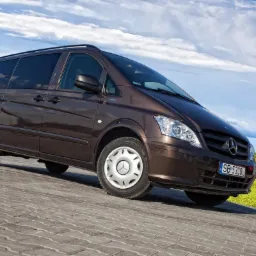 Brązowy Mercedes-Benz Vito, widok z przodu pod kątem, na tle błękitnego nieba z chmurami i zielonego pola w oddali.