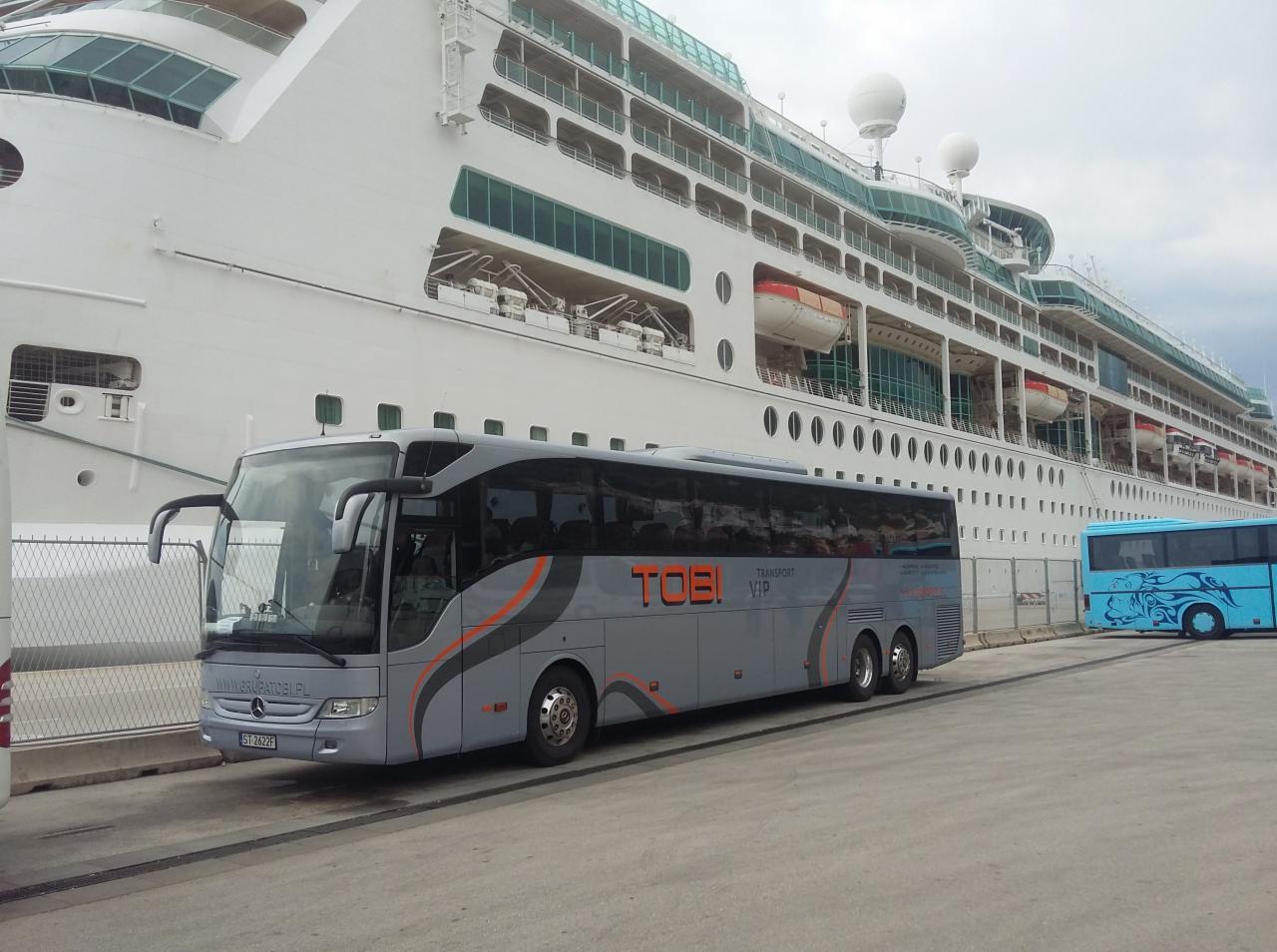 Szary autokar z pomarańczowymi akcentami marki TOBI VIP zaparkowany przed ogromnym białym statkiem wycieczkowym, z widocznym fragmentem innego, niebieskiego autokaru w tle.