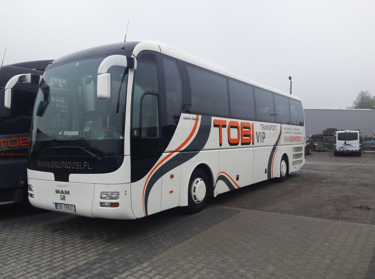 Biały autokar z czarno-pomarańczowymi akcentami i napisem TOBI VIP Transport na tle szarej, brukowej nawierzchni i innych pojazdów w tle.