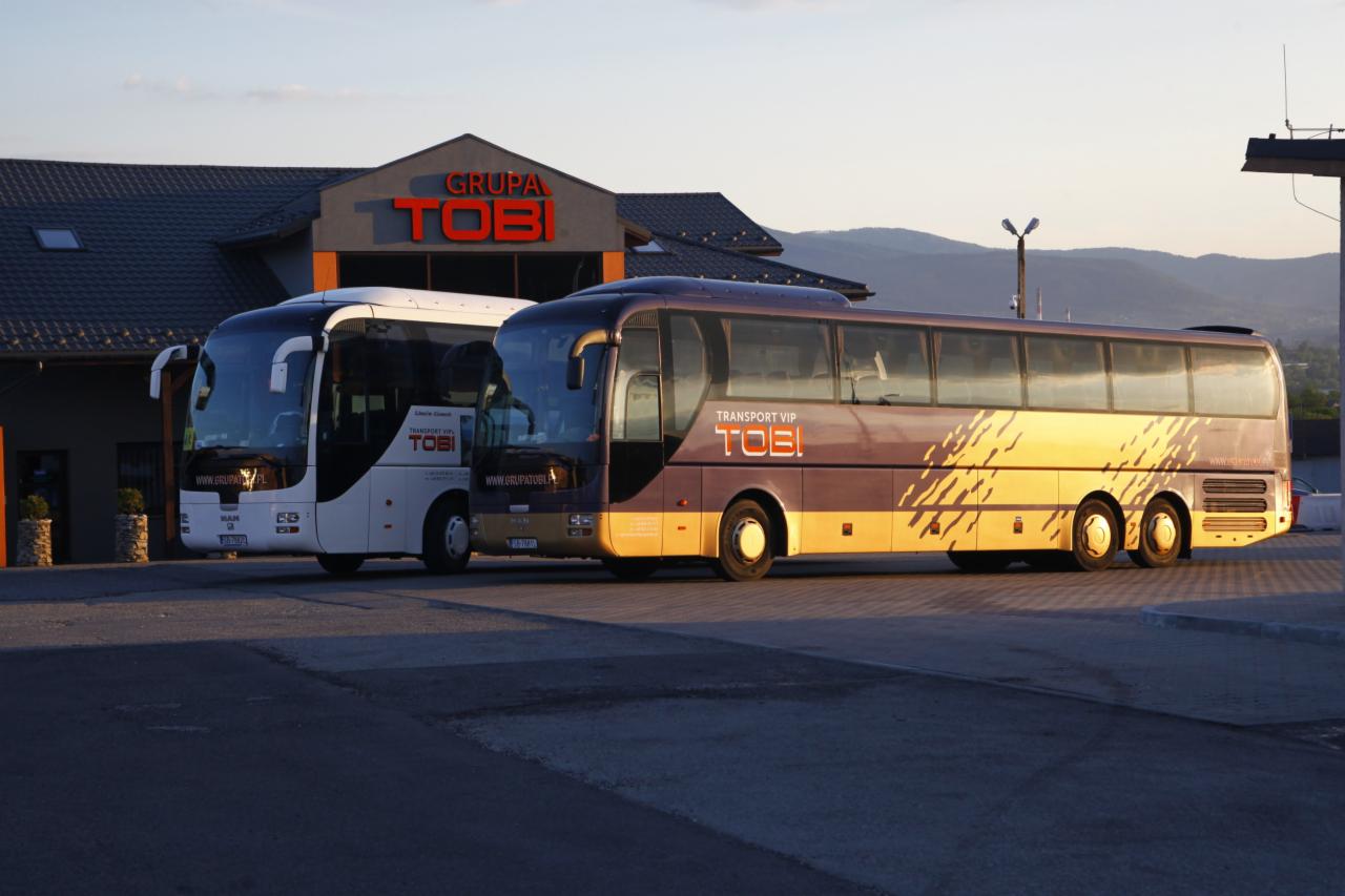 Dwa autokary firmy transportowej TOBI zaparkowane przed budynkiem firmy, jeden biały, drugi bordowo-złoty z napisem TRANSPORT VIP, w tle góry i błękitne niebo o zachodzie słońca.