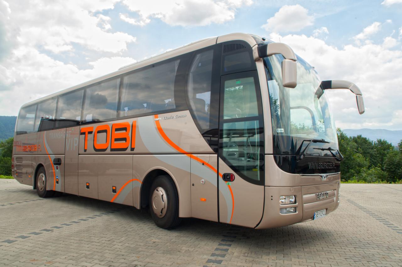 Autokar turystyczny TOBI VIP Lion's Coach w kolorze beżowym z pomarańczowymi akcentami, zaparkowany na brukowanym placu z widokiem na zalesione wzgórza i pochmurne niebo.