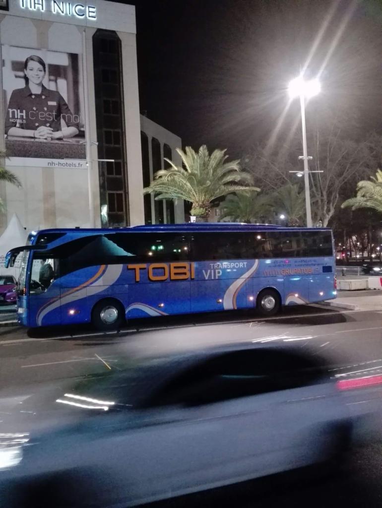 Niebieski autokar TOBI VIP zaparkowany nocą przed budynkiem NH Nice, widoczna reklama hotelu i palmy.