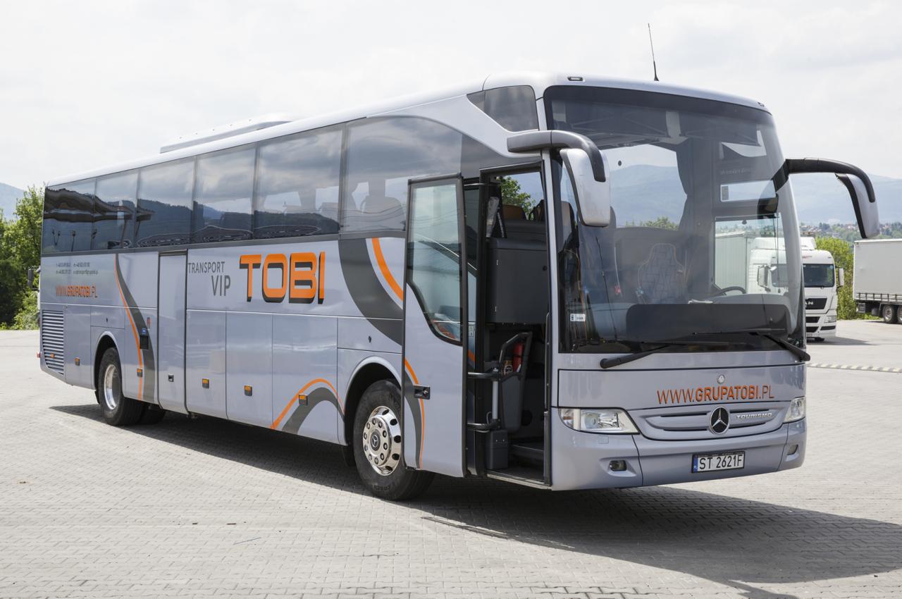 Nowoczesny, srebrny autokar z otwartymi drzwiami, widoczny z boku, z logo firmy 'TOBI' i adresem strony internetowej, zaparkowany na parkingu z widokiem na góry w tle.