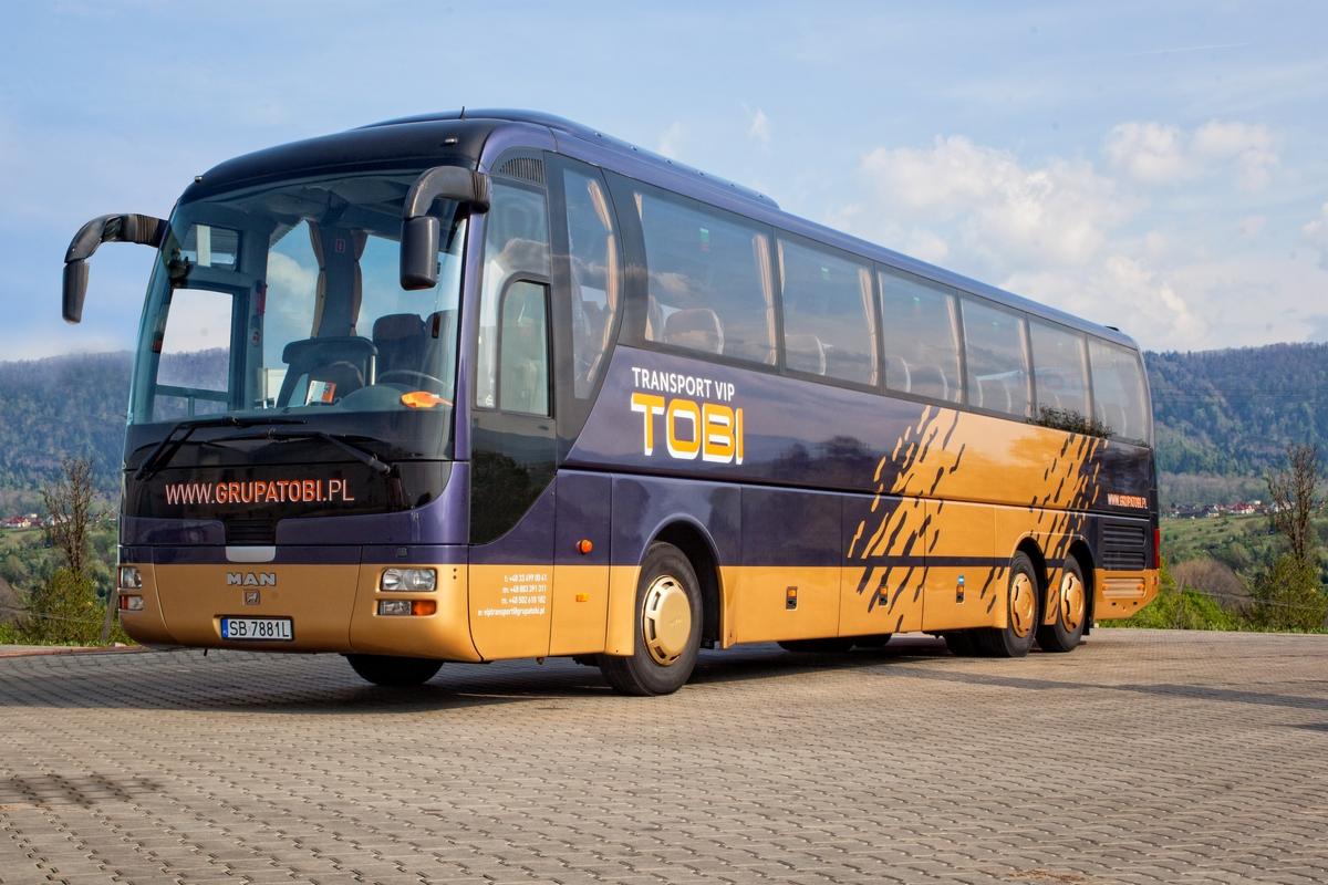 Autokar turystyczny w kolorach fioletowo-złotych z napisem 'TRANSPORT VIP TOBI', zaparkowany na brukowanej powierzchni z widokiem na góry w tle.