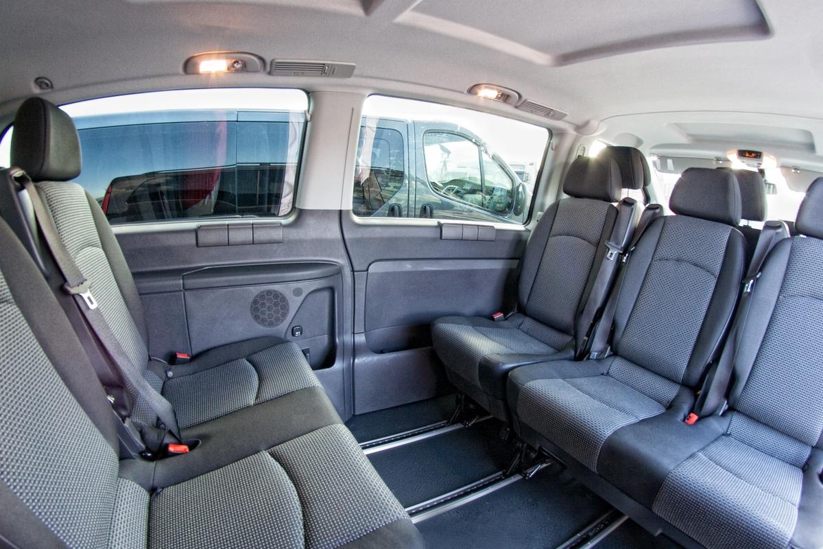 Wnętrze przestronnego busa pasażerskiego z trzema rzędami foteli obitych szarą tkaniną w drobną kratkę, widoczne przesuwne drzwi i szyny mocujące fotele w podłodze.