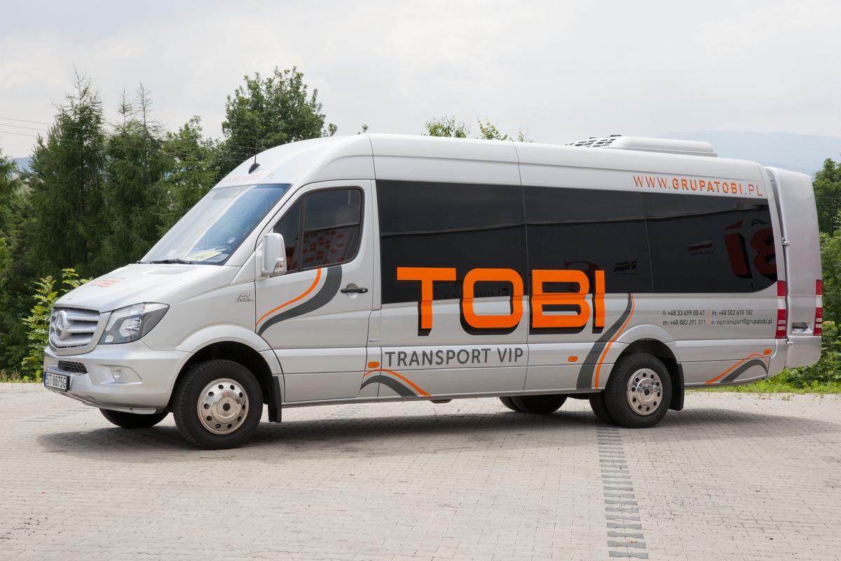 Srebrny bus Mercedes-Benz z przyciemnianymi szybami, oklejony pomarańczowo-szarymi grafikami i napisem TOBI TRANSPORT VIP, zaparkowany na betonowej nawierzchni, w tle drzewa.