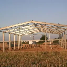 Szkielet powstającej hali stalowej na tle zaoranego pola i błękitnego nieba, widoczne spawy i elementy konstrukcyjne.