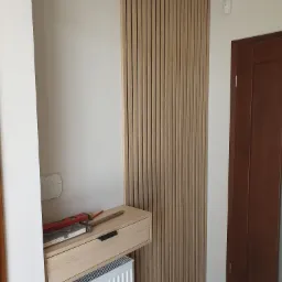 Drewniana ścianka działowa z pionowych listew i szuflada zawieszona nad grzejnikiem, na blacie poziomica, ołówek i notatnik