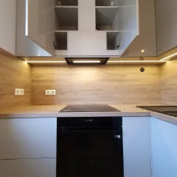 Nowoczesna kuchnia z szarymi szafkami i drewnianym blatem, oświetleniem LED pod szafkami wiszącymi, otwartymi szafkami górnymi i czarną płytą indukcyjną z piekarnikiem.