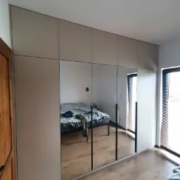 Beżowa szafa wnękowa z lustrzanymi drzwiami przesuwnymi, odbijająca fragment sypialni z łóżkiem i oknem.